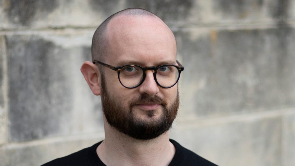 Headshot of TJ Robinson, wearing thick-rimmed glasses and a black crewneck sweater, standing in front of a concrete wall.